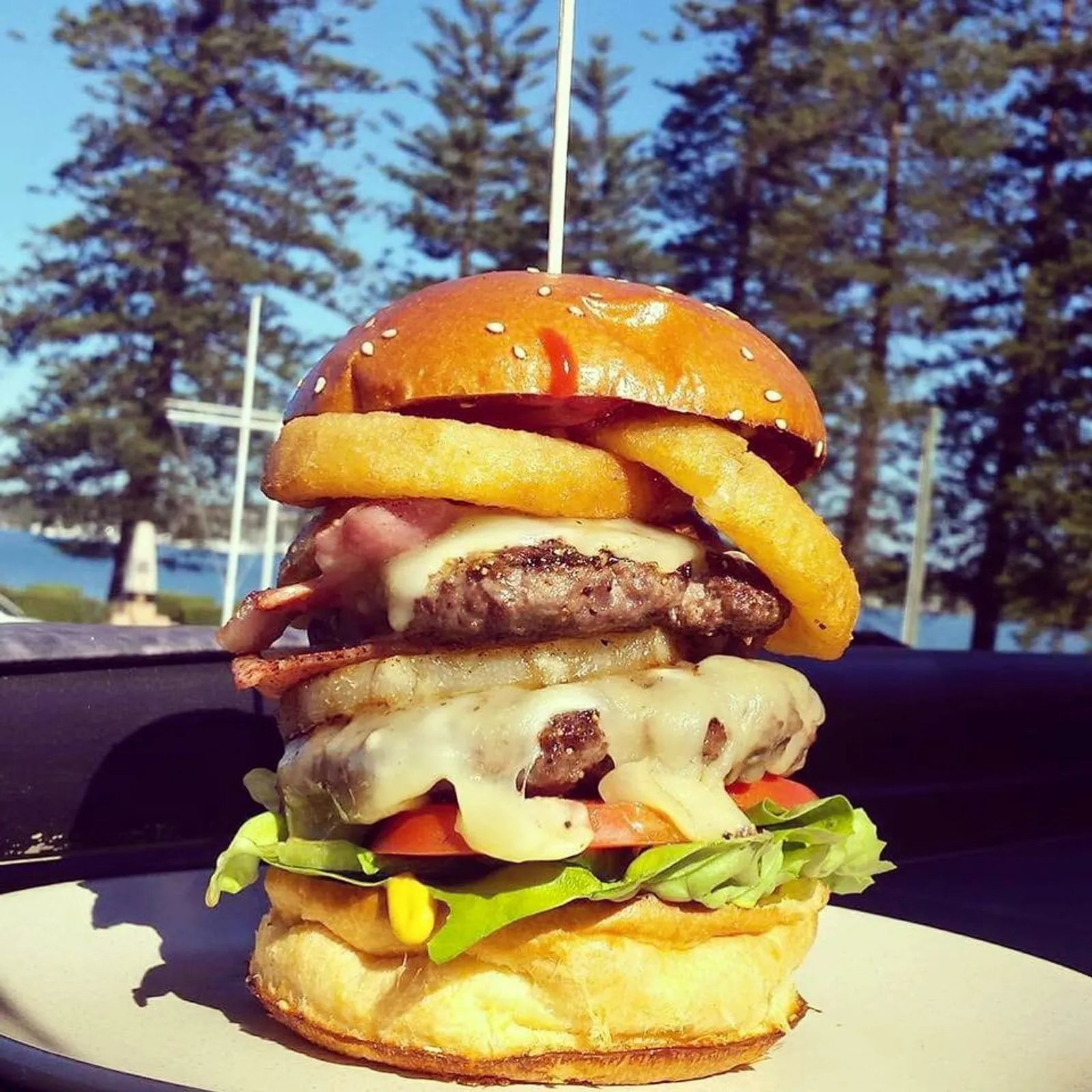 Towering double-patty burger with cheese, bacon and onion rings against coastal pine backdrop