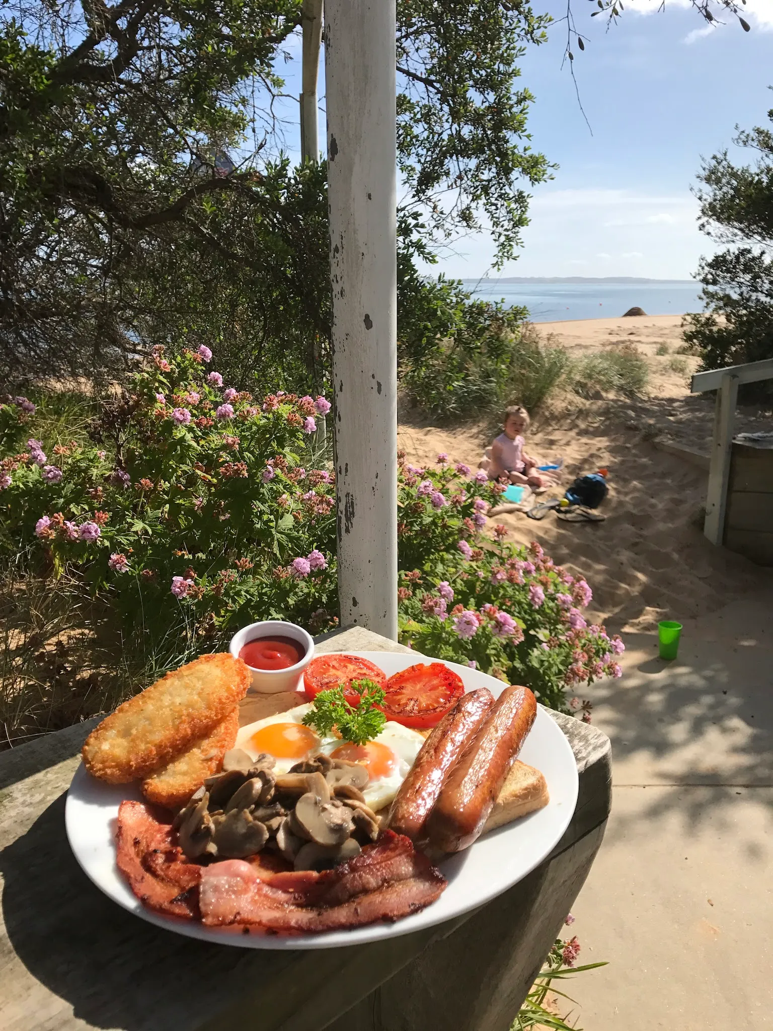 Full cooked breakfast plate outdoors with flowering dunes, beach and blue bay behind