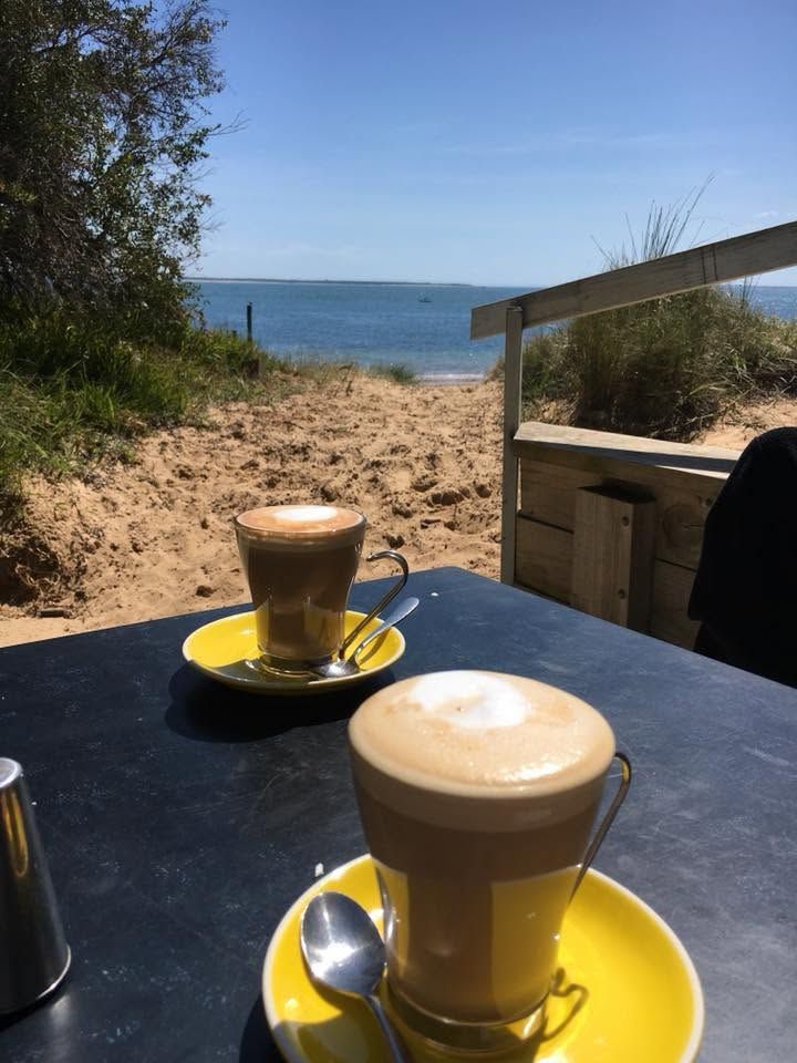 Two lattes in glass cups on yellow saucers with sandy beach and blue bay in the background